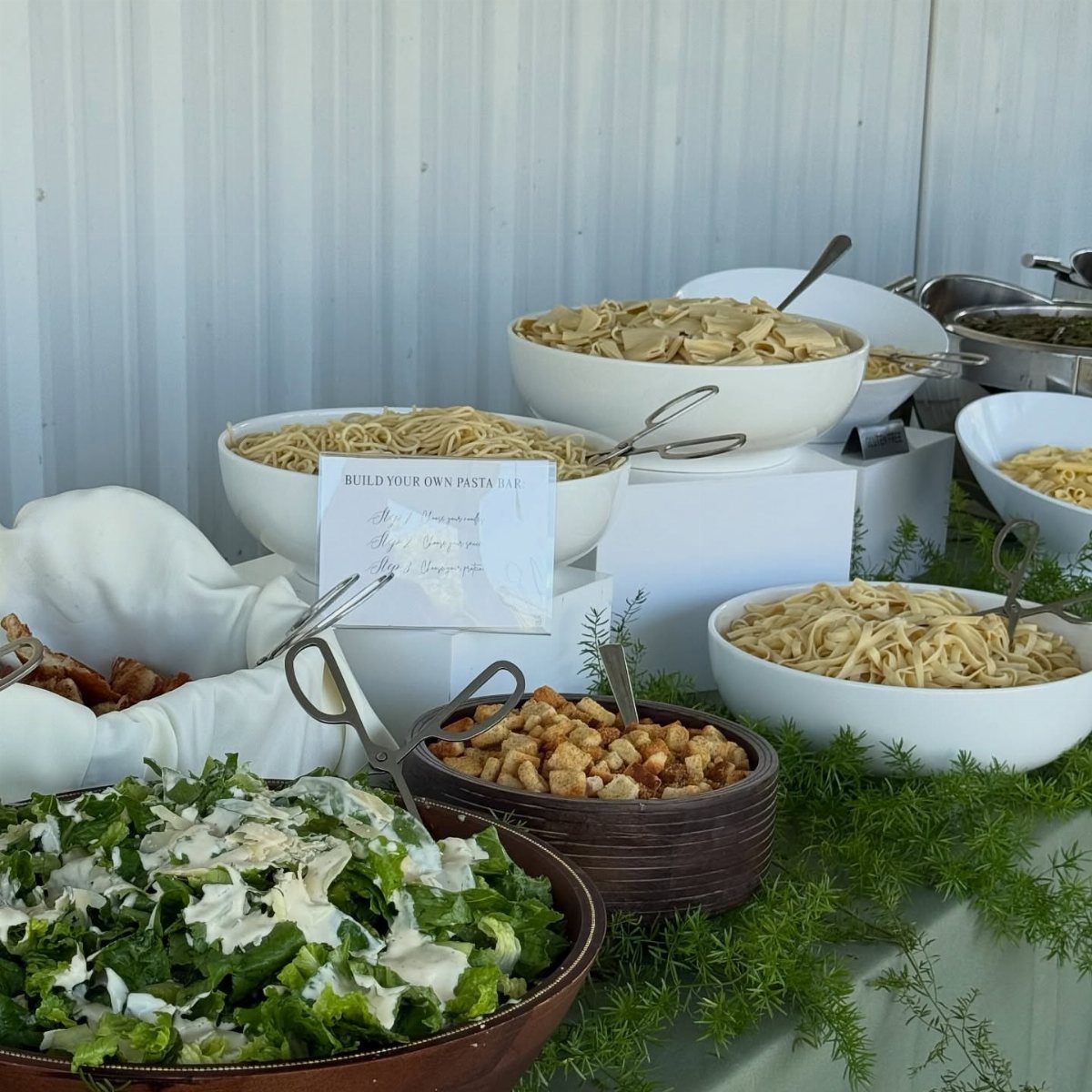 Pasta buffet station with various noodles, salad, and croutons on white pedestals.