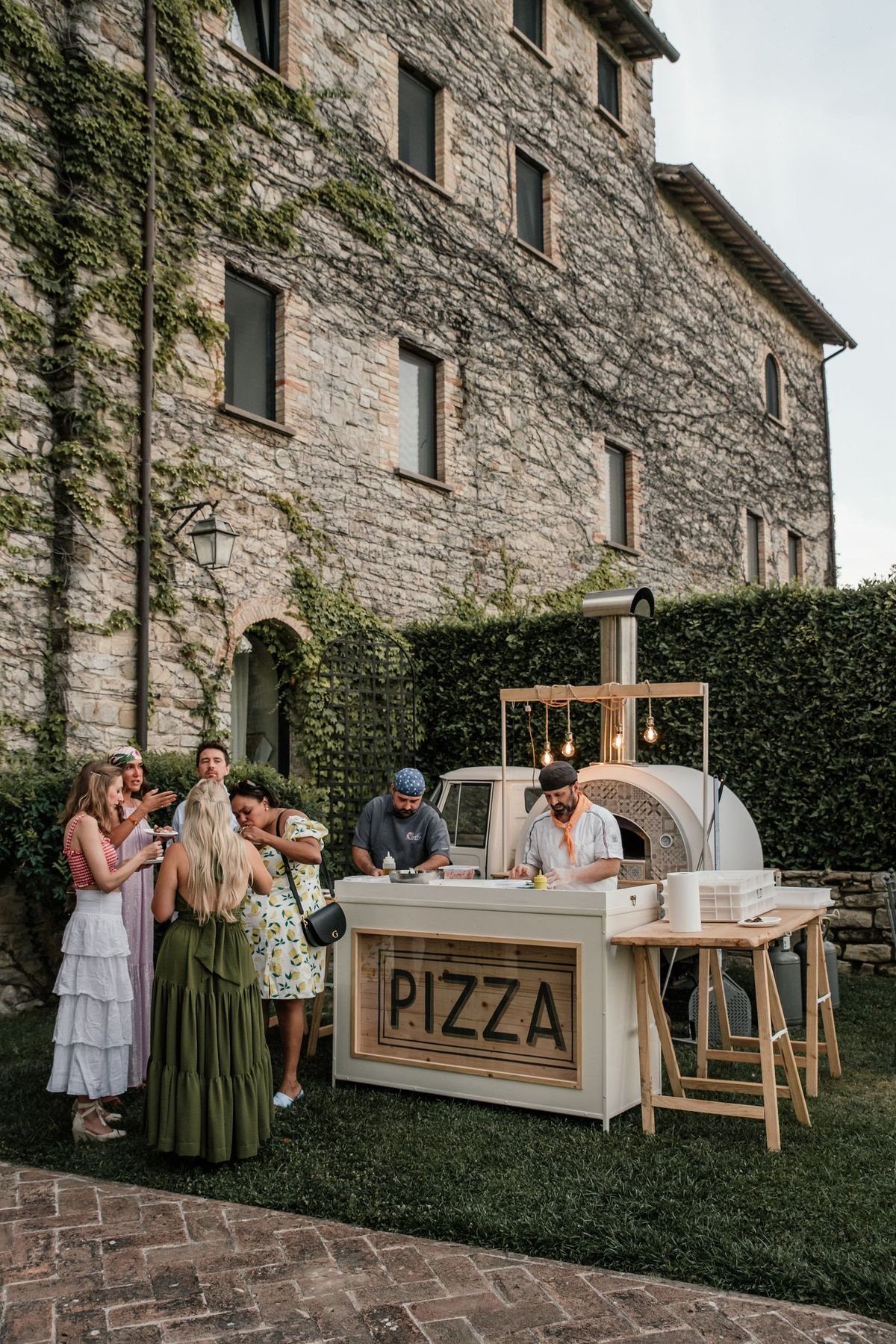 Outdoor pizza station with a wood-fired oven and guests at a rustic venue.