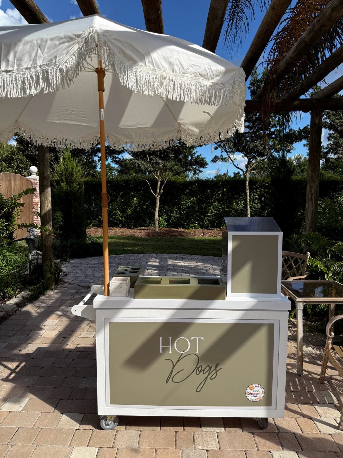 Hot dog cart with a white fringed umbrella on a brick patio.