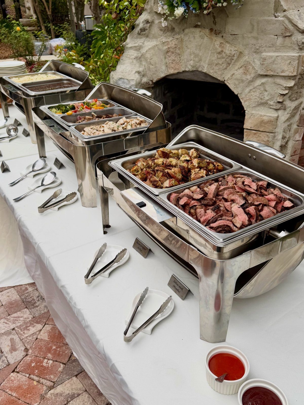 Buffet table with sliced grilled meats and roasted vegetables in stainless steel chafing dishes.