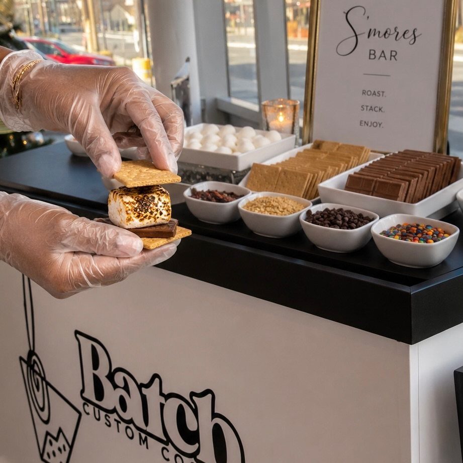 Hands assembling a s'more at a dessert station with toppings and chocolate.