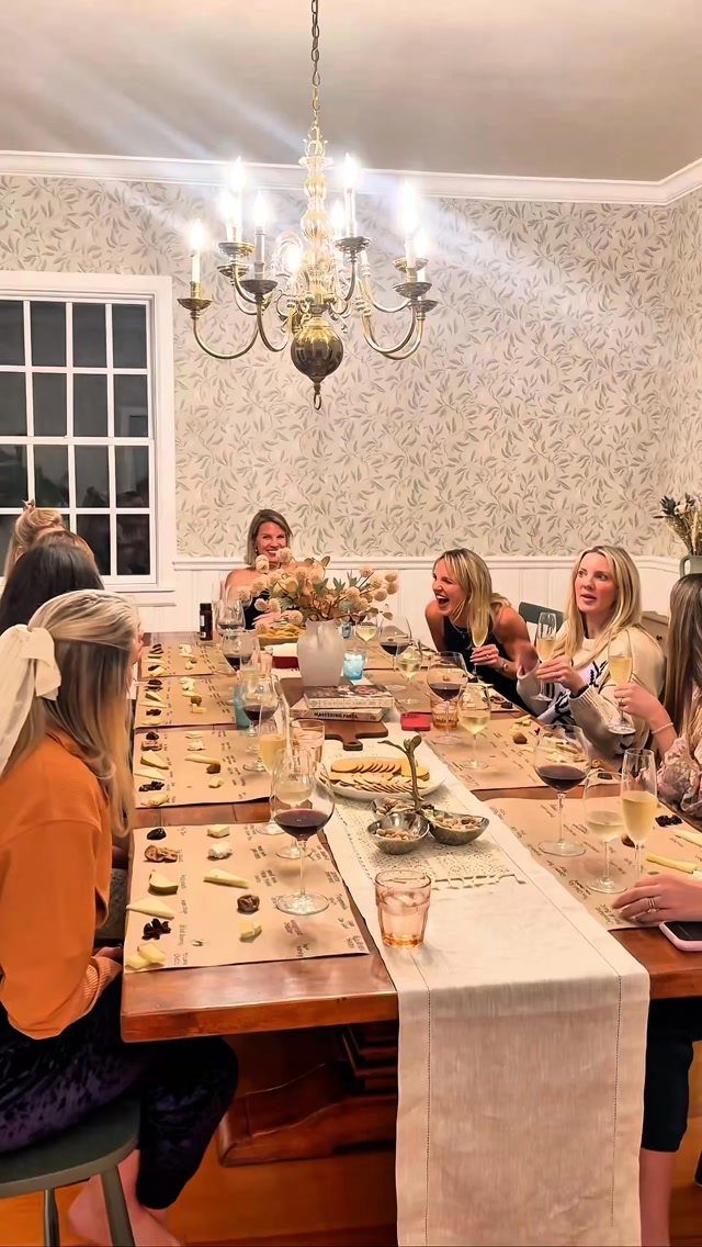 Women enjoying wine and appetizers at a long dining table under a chandelier.