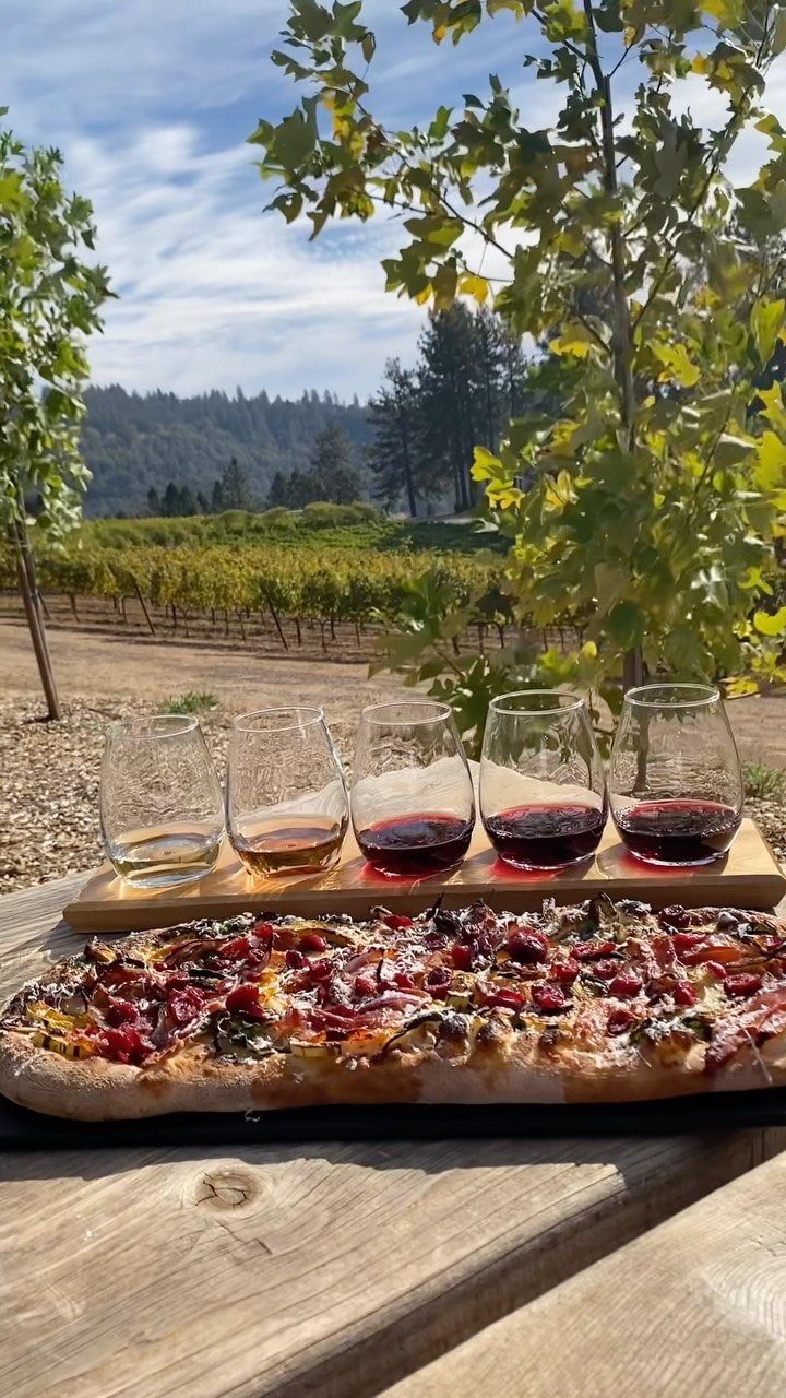 Wine glasses and flatbread pizza on a wooden table overlooking a sunny vineyard.