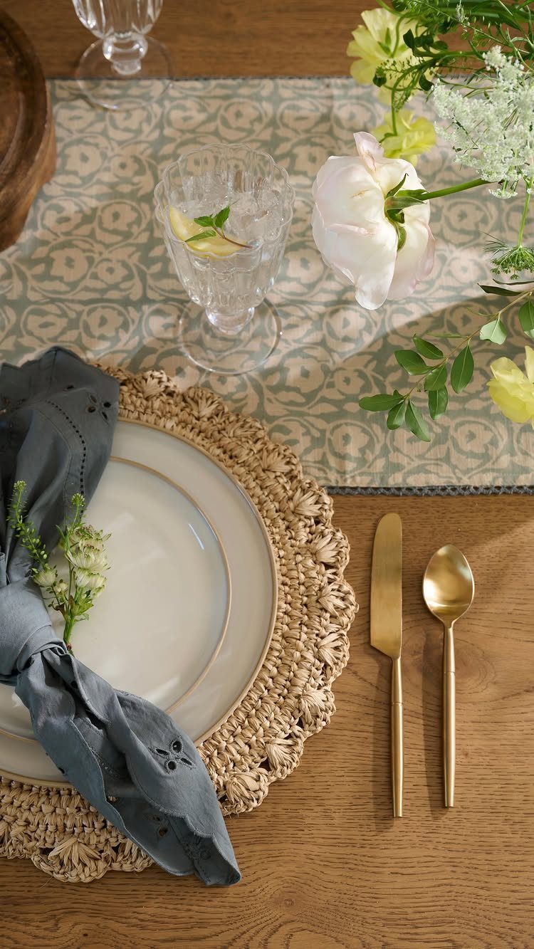 Wooden table setting with white plate, woven charger, blue napkin, and gold cutlery.