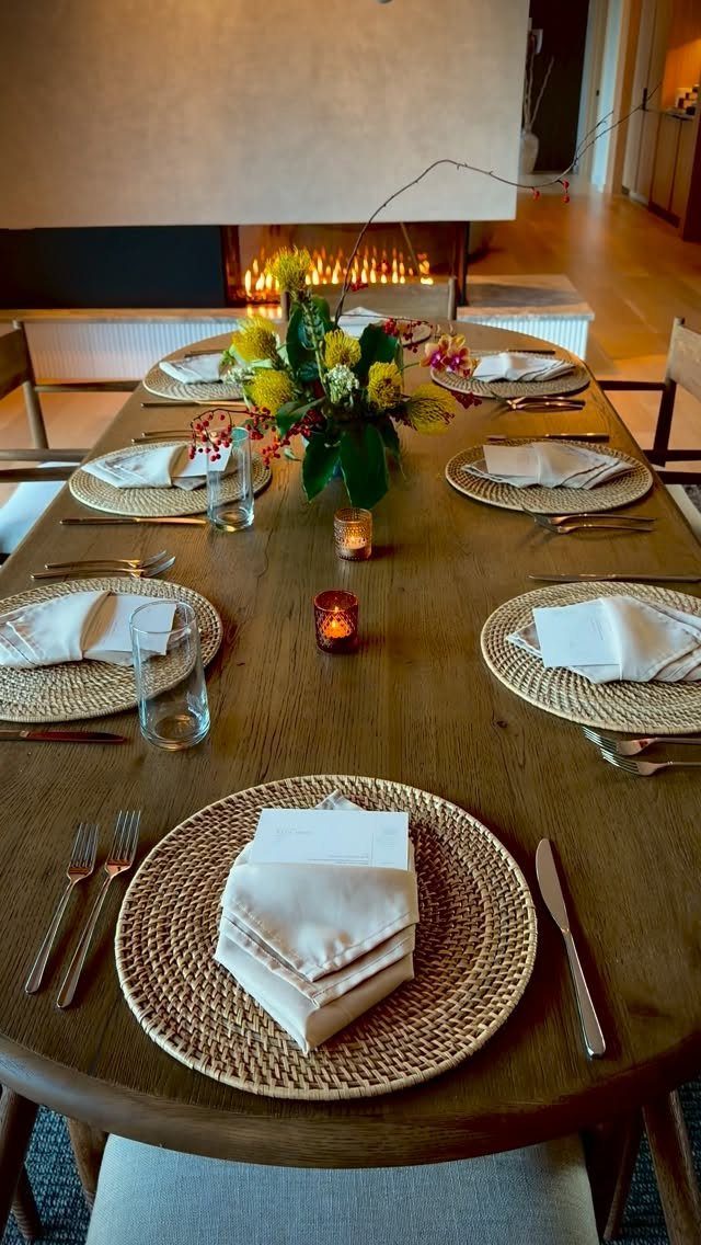 Wooden dining table set with woven placemats, white napkins, and a floral centerpiece.